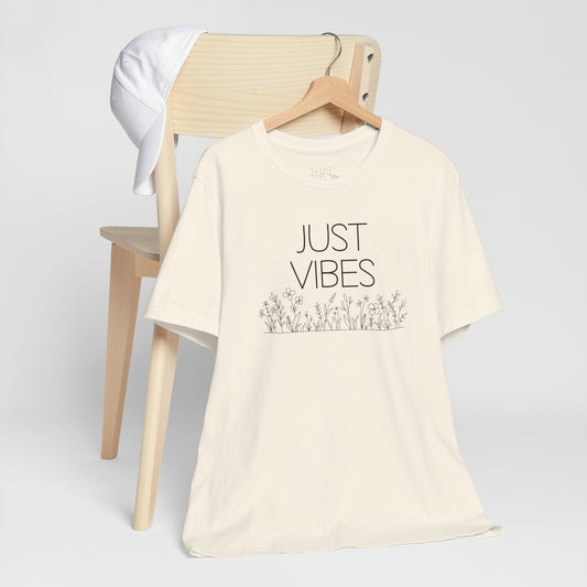 Just Vibes Graphic T-Shirt in cream color with minimalist floral design displayed on a wooden chair with a white cap.