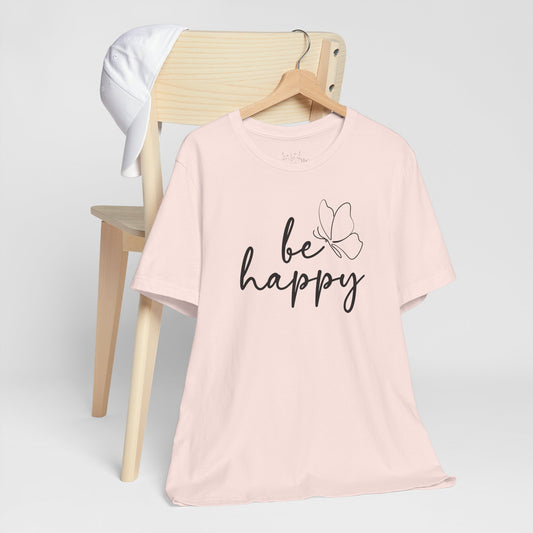 Be Happy graphic t-shirt in soft pink with minimalist butterfly design displayed on a wooden chair with a white cap for a chic look.
