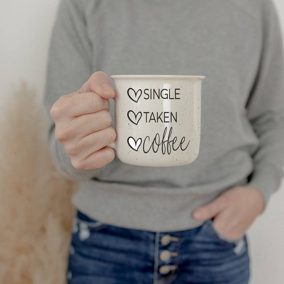 White ceramic mug with 'Single Taken Coffee' text, held by a person wearing a gray sweater and jeans in a casual setting.