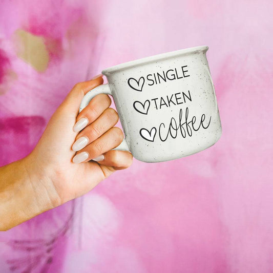White ceramic mug with 'Single Taken Coffee' text, held by a hand with manicured nails, set against a pink floral background.