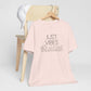 Just Vibes Graphic T-Shirt in light pink with minimalist floral design displayed on a wooden chair with a white cap.