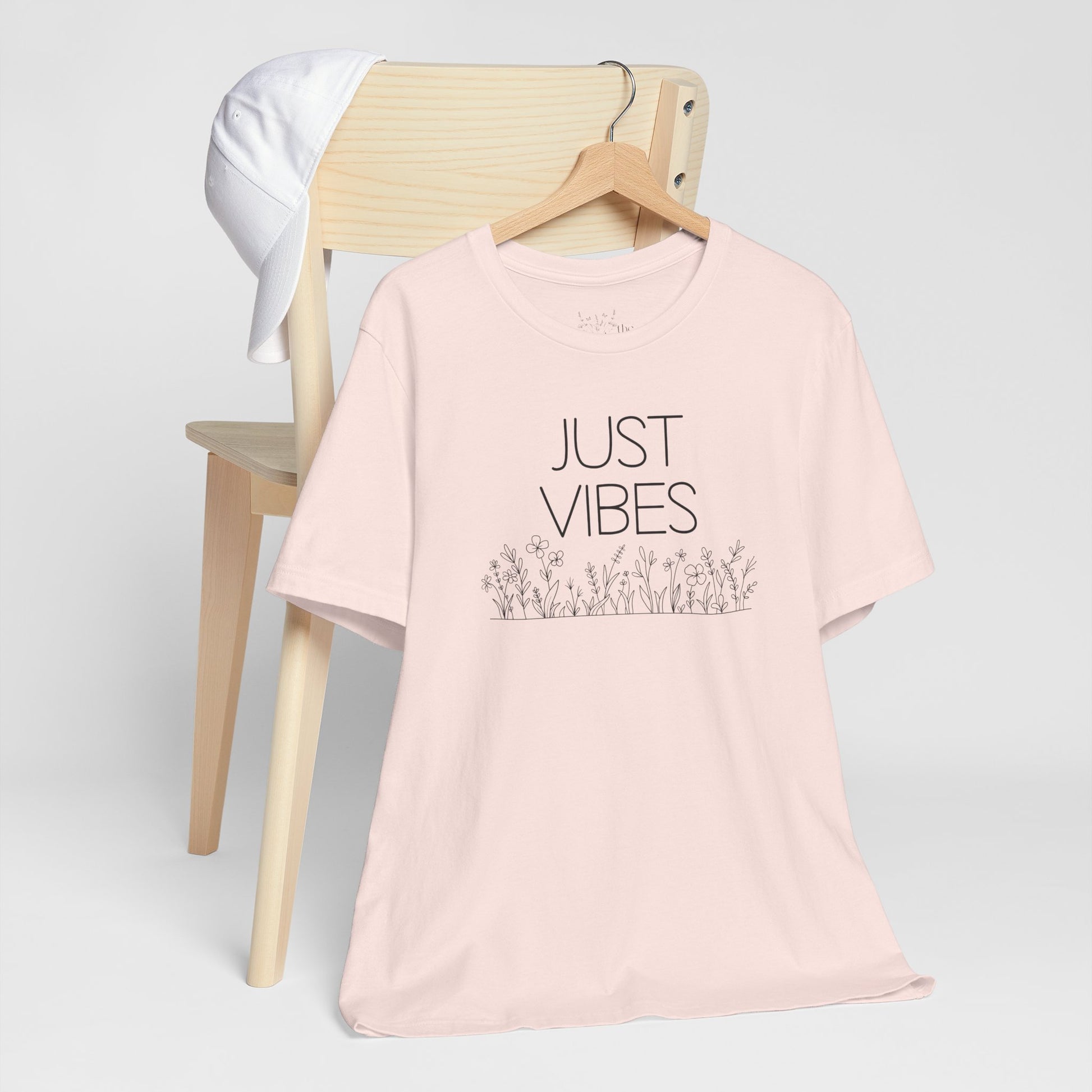 Just Vibes Graphic T-Shirt in light pink with minimalist floral design displayed on a wooden chair with a white cap.