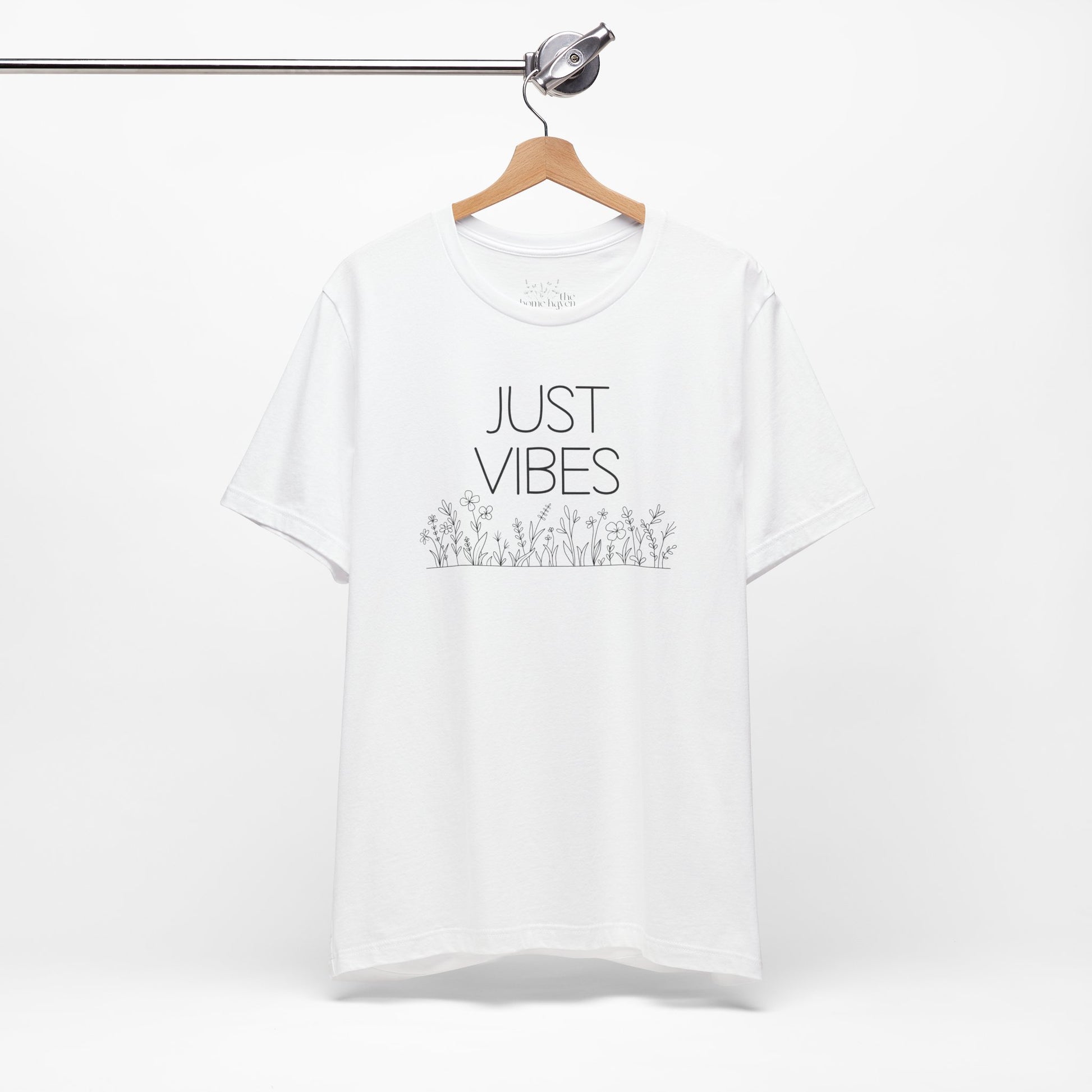 Just Vibes Graphic T-Shirt in white with minimalist floral design hanging on a wooden hanger against a white background.