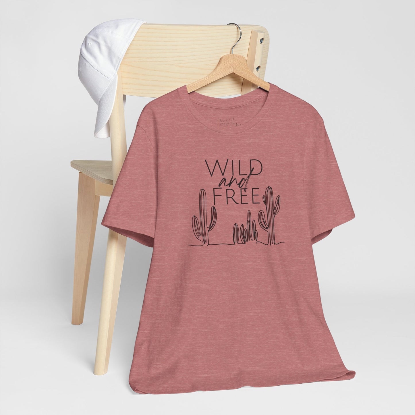 Rust-colored Wild and Free cactus graphic T-shirt draped over a chair, paired with a white cap for a casual and trendy vibe.