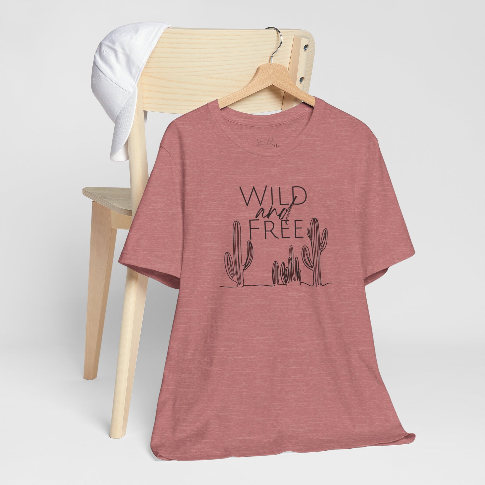 Rust-colored Wild and Free cactus graphic T-shirt draped over a chair, paired with a white cap for a casual and trendy vibe.
