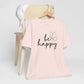 Be Happy graphic t-shirt in soft pink with minimalist butterfly design displayed on a wooden chair with a white cap for a chic look.