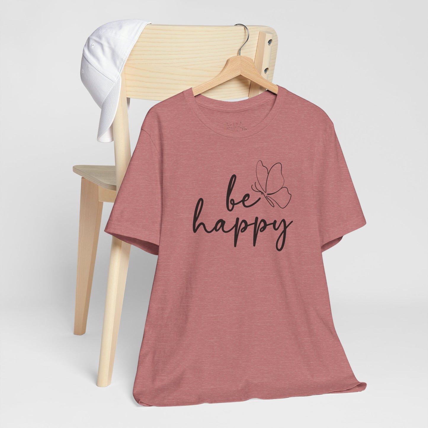 Be Happy graphic t-shirt in dusty rose with minimalist butterfly design displayed on a wooden chair with a white cap for a relaxed look.