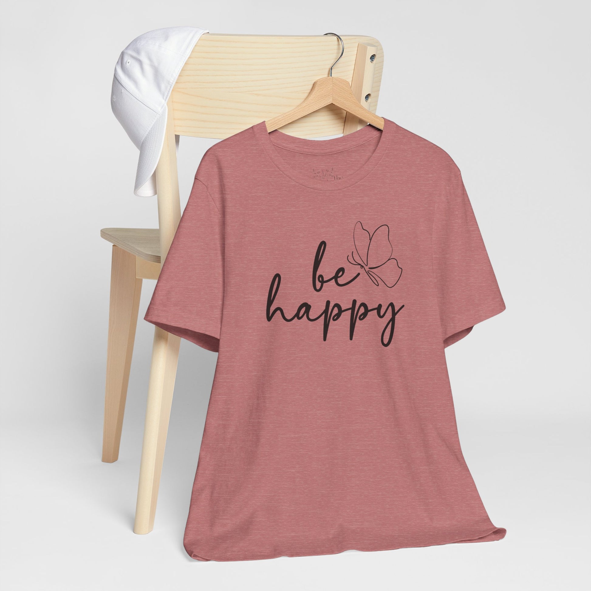 Be Happy graphic t-shirt in dusty rose with minimalist butterfly design displayed on a wooden chair with a white cap for a relaxed look.