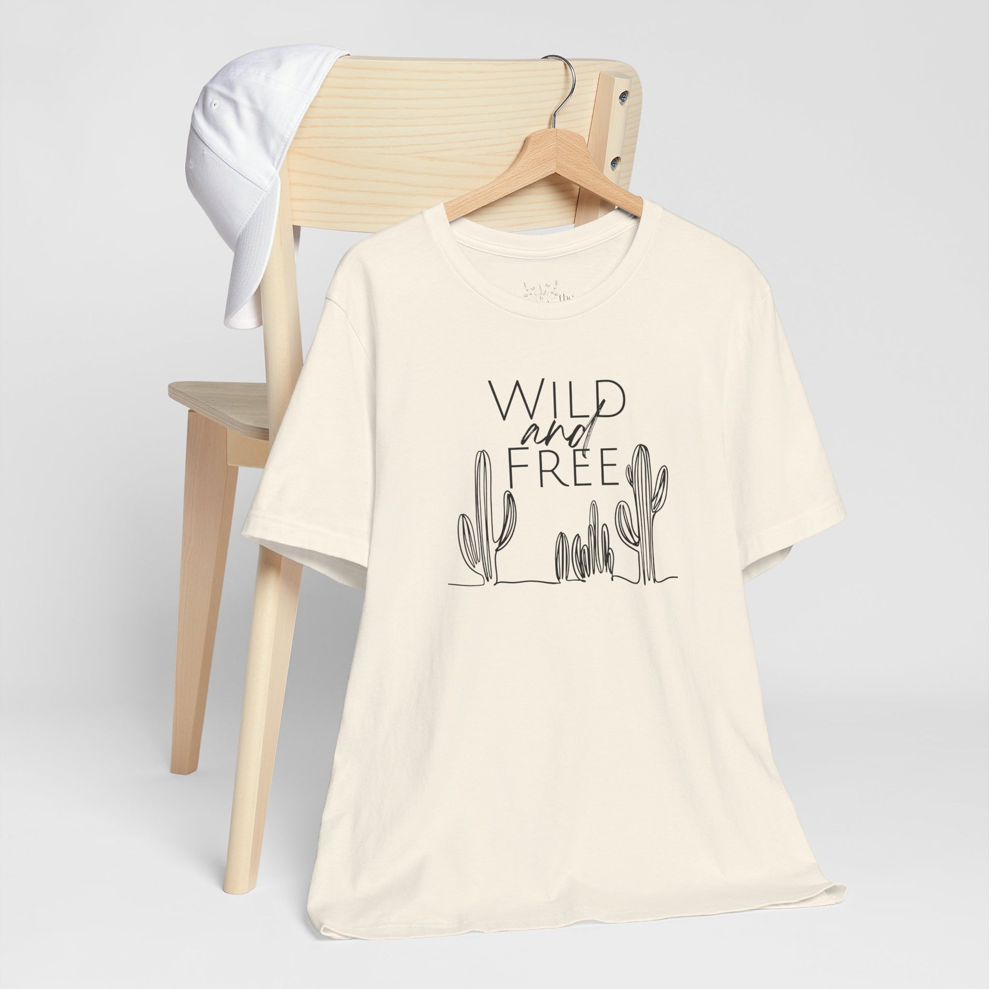 Cream Wild and Free cactus graphic T-shirt draped over a chair, styled with a white cap for a casual and nature-inspired outfit.