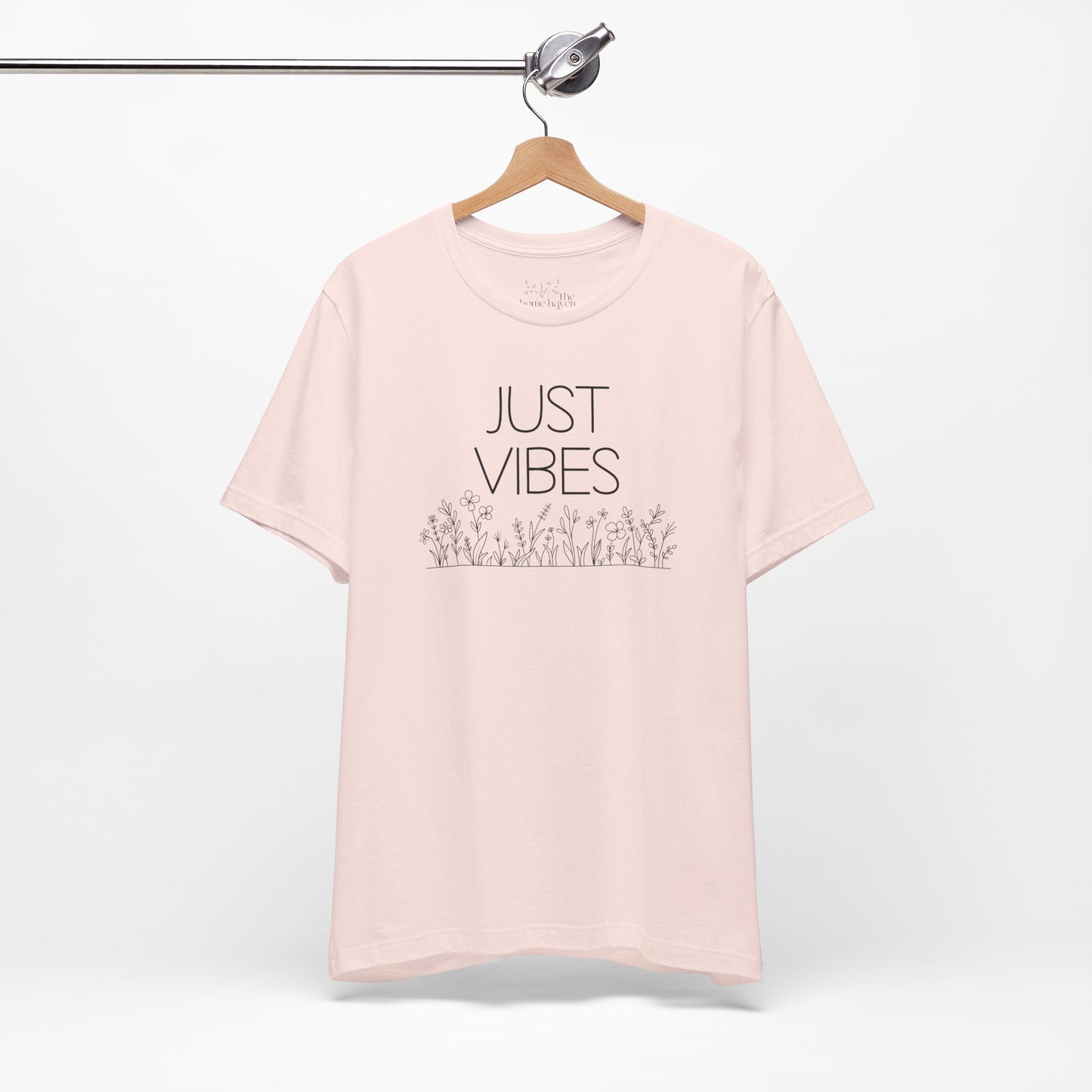 Just Vibes Graphic T-Shirt in light pink with minimalist floral design hanging on a wooden hanger against a white background.