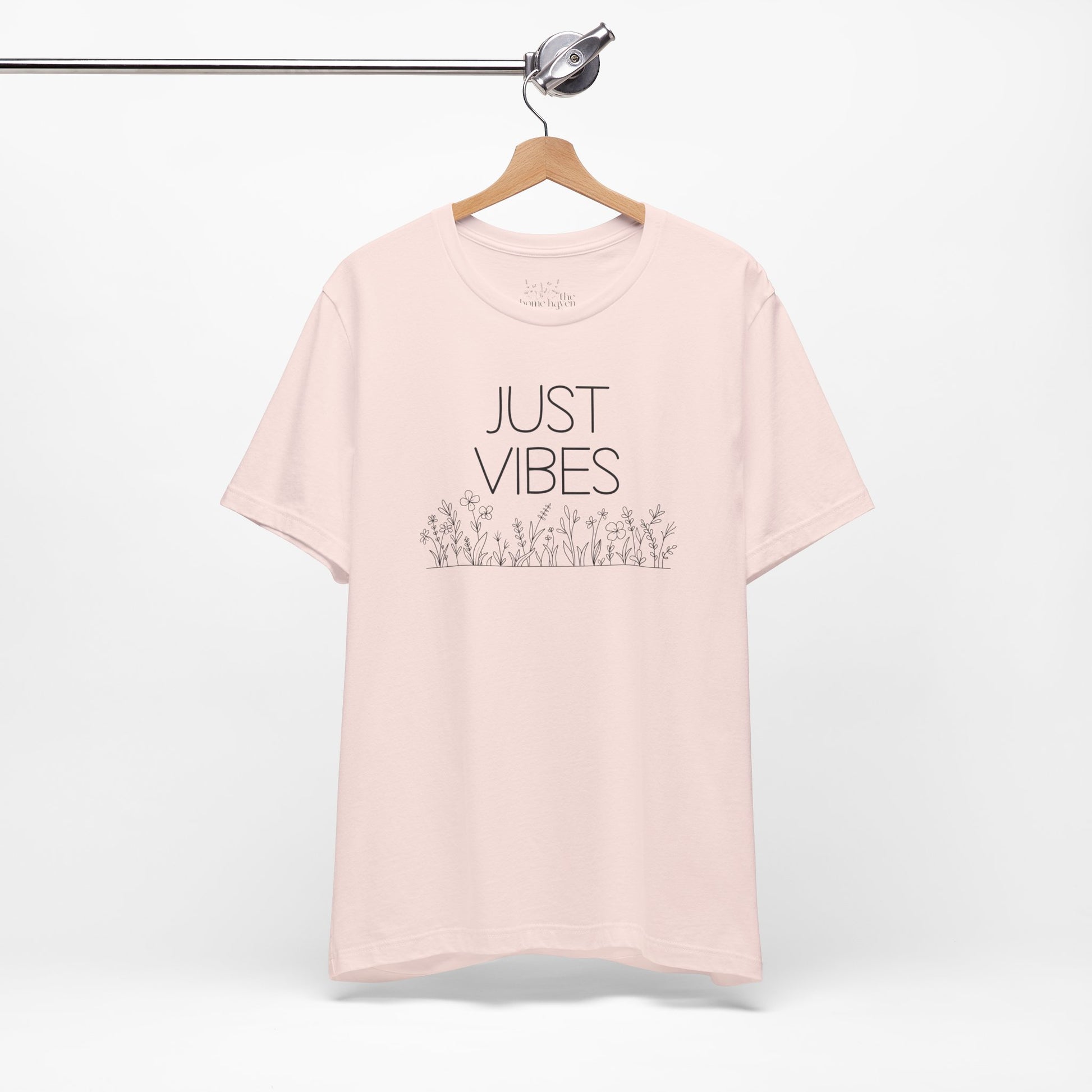 Just Vibes Graphic T-Shirt in light pink with minimalist floral design hanging on a wooden hanger against a white background.
