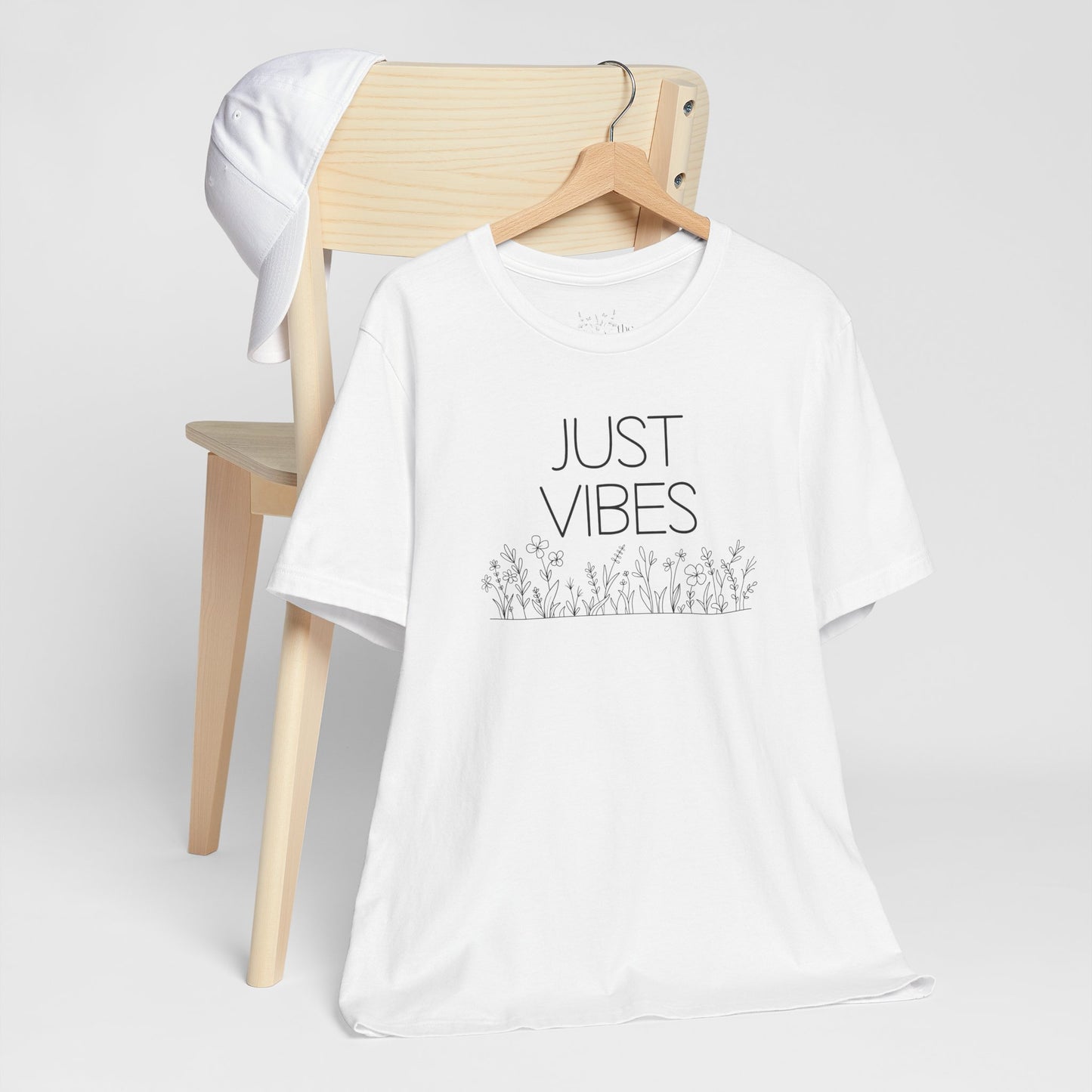 Just Vibes Graphic T-Shirt in white with minimalist floral design displayed on a wooden chair with a white cap.
