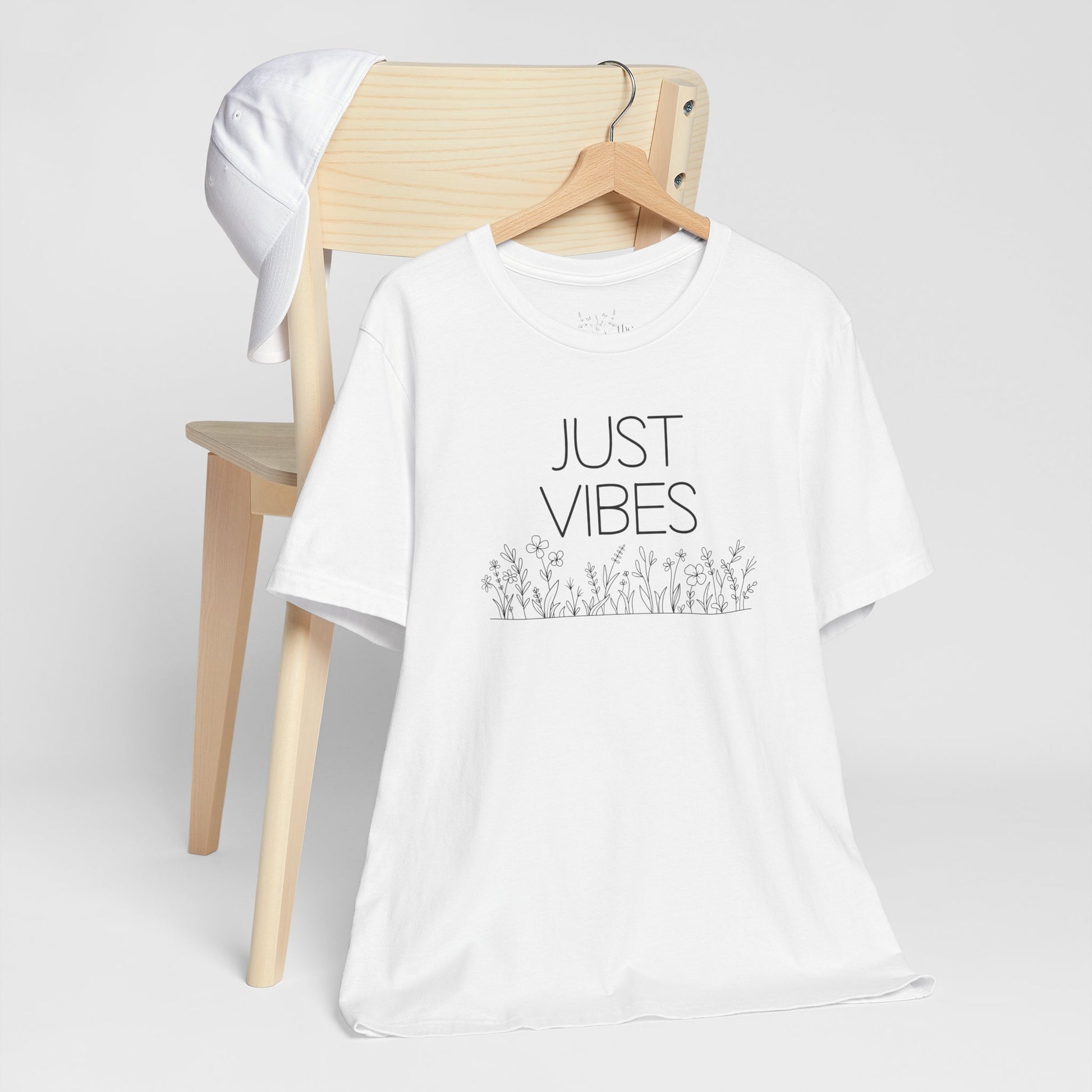 Just Vibes Graphic T-Shirt in white with minimalist floral design displayed on a wooden chair with a white cap.