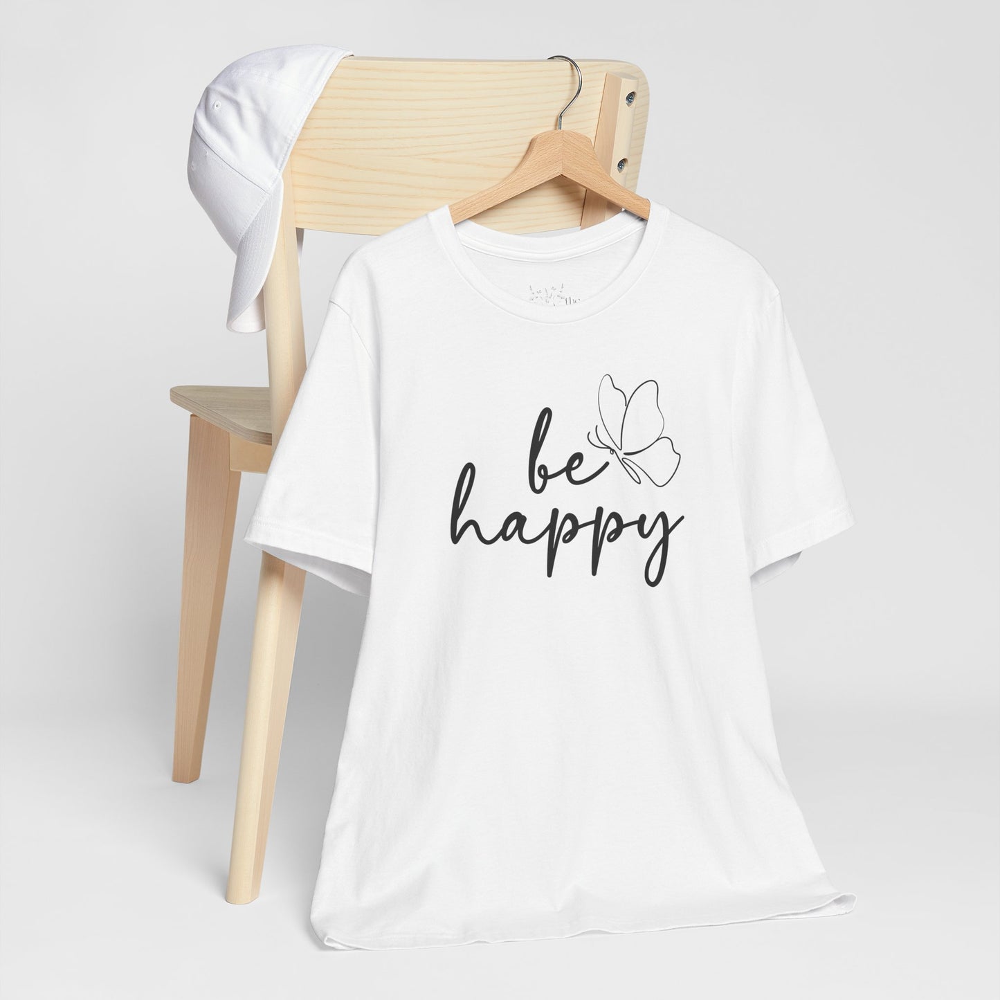 White Be Happy graphic t-shirt with minimalist butterfly design displayed on a wooden chair with a white cap for a fresh style.