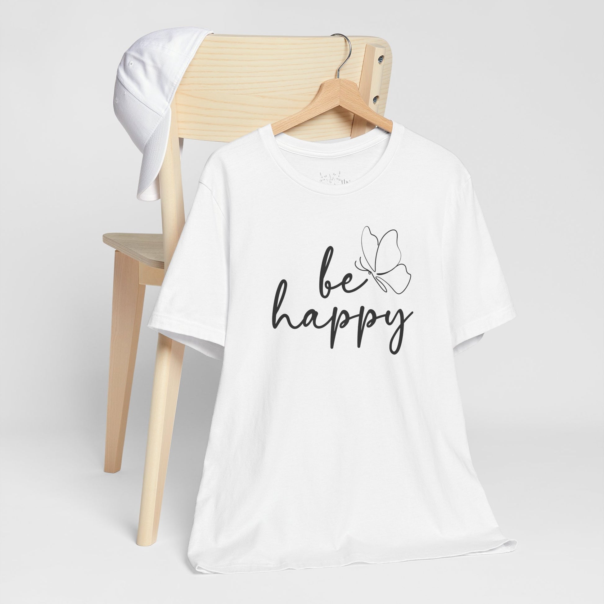 White Be Happy graphic t-shirt with minimalist butterfly design displayed on a wooden chair with a white cap for a fresh style.