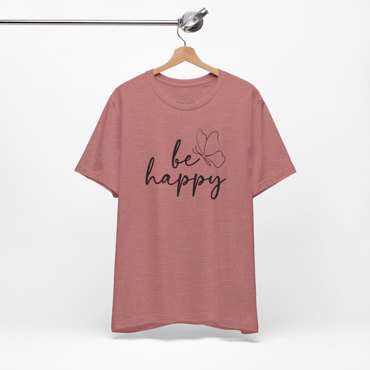 Be Happy graphic t-shirt in dusty rose with minimalist butterfly design hanging on a wooden hanger against a white background.