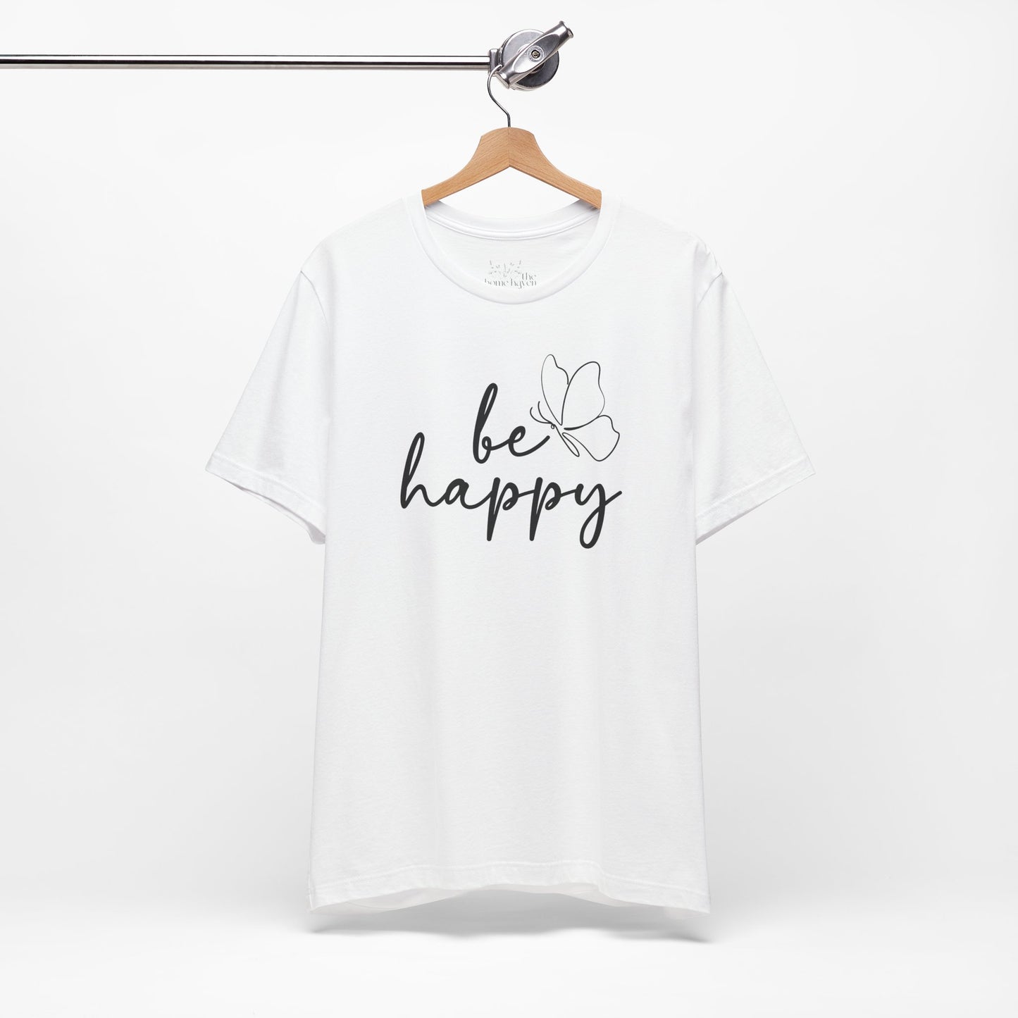 White Be Happy graphic t-shirt with minimalist butterfly design hanging on a wooden hanger for a clean and modern presentation.