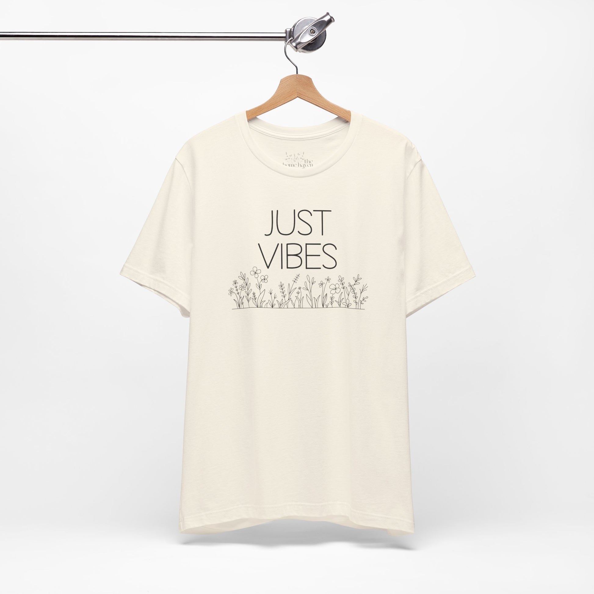 Just Vibes Graphic T-Shirt in cream color with minimalist floral design hanging on a wooden hanger against a white background.