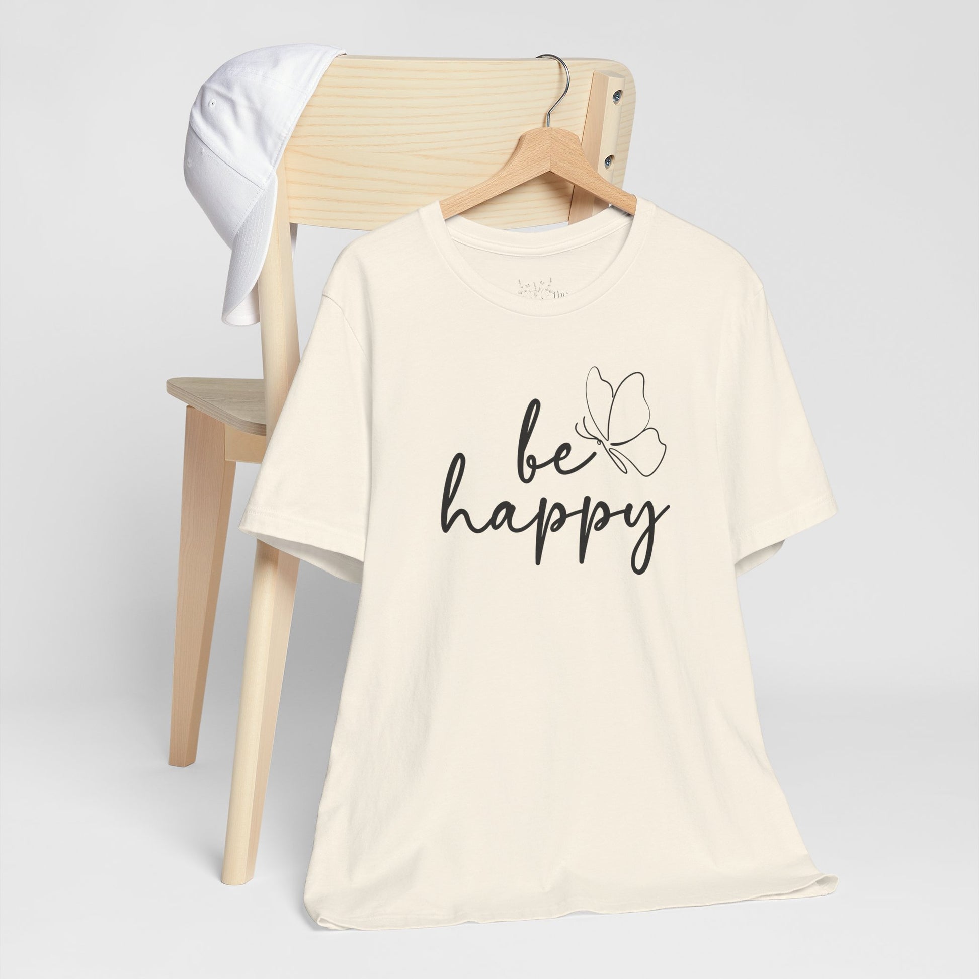 Be Happy graphic t-shirt in cream with minimalist butterfly design displayed on a wooden chair with a white cap for a cozy look.