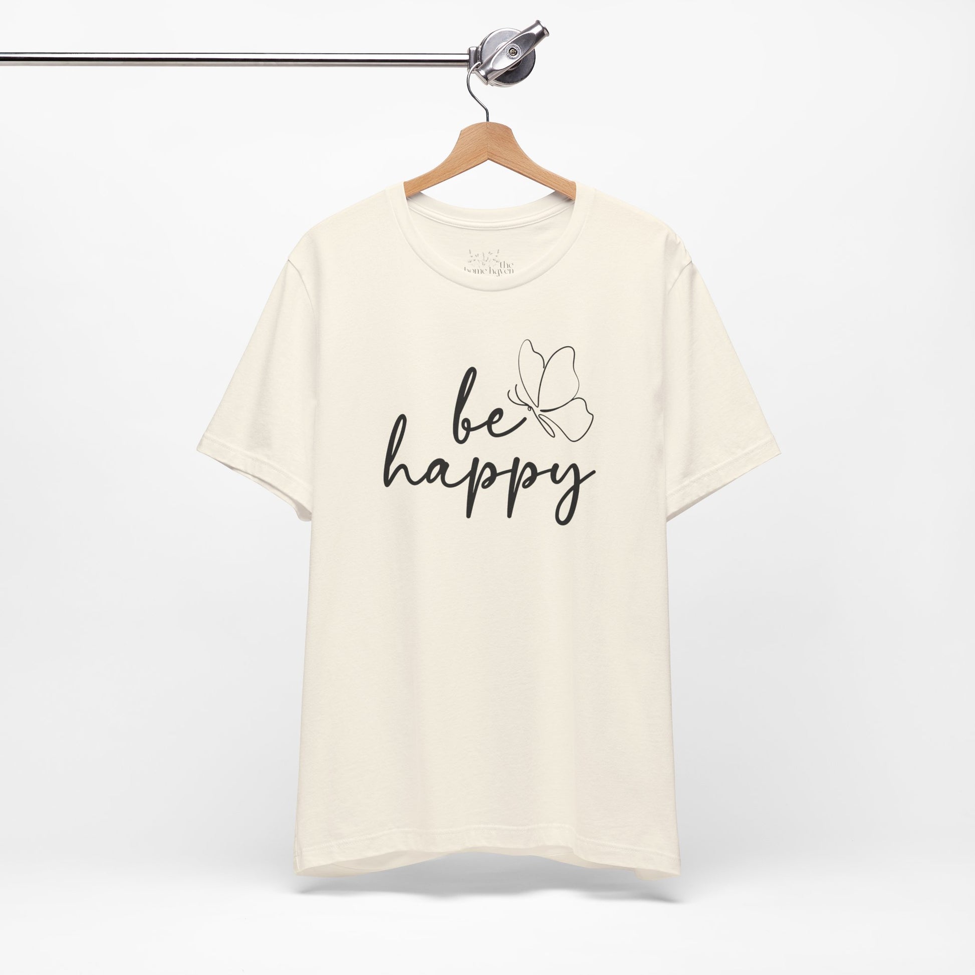 Cream Be Happy graphic t-shirt with minimalist butterfly design hanging on a wooden hanger, perfect for casual and chic outfits.