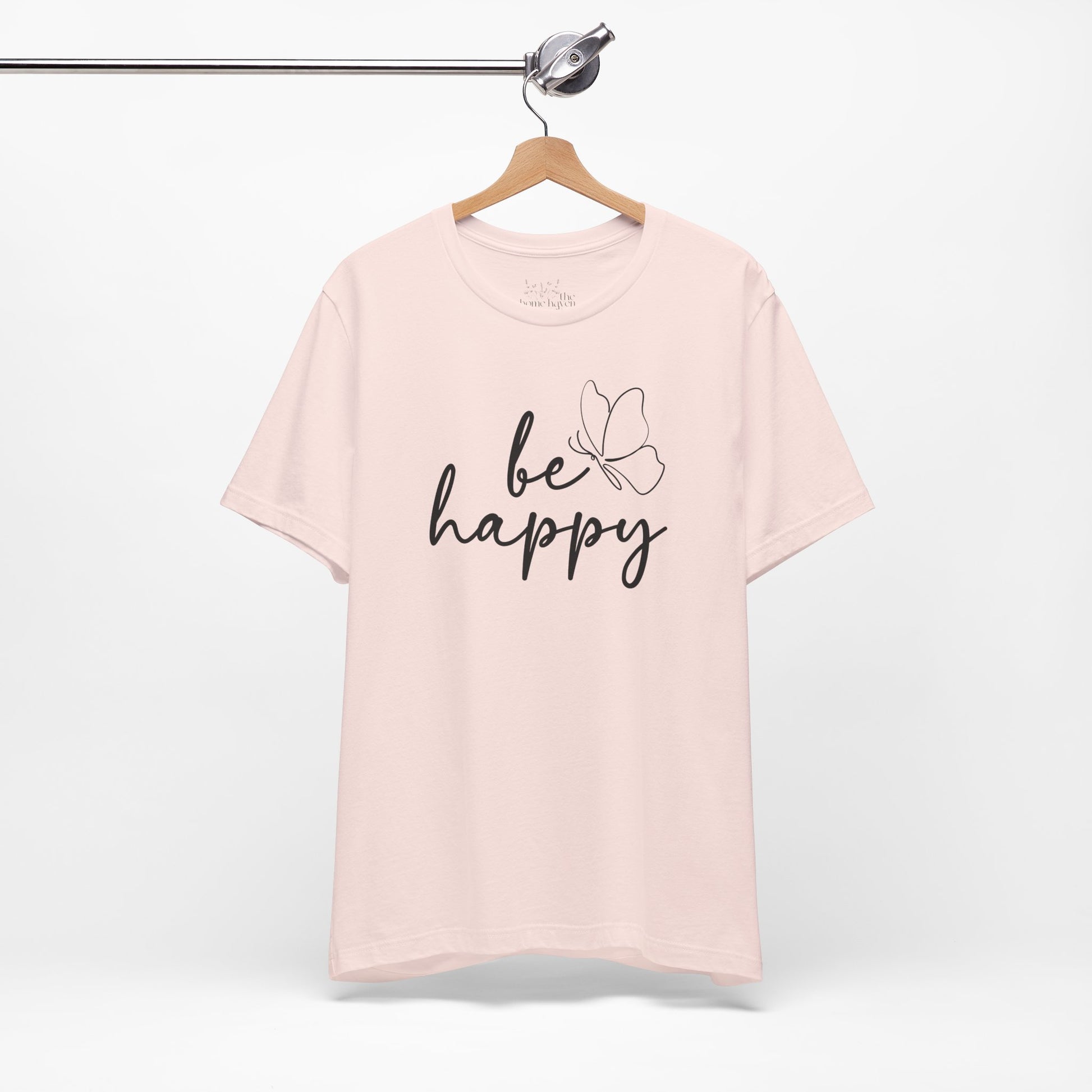 Be Happy graphic t-shirt in soft pink with minimalist butterfly design hanging on a wooden hanger for a clean display.