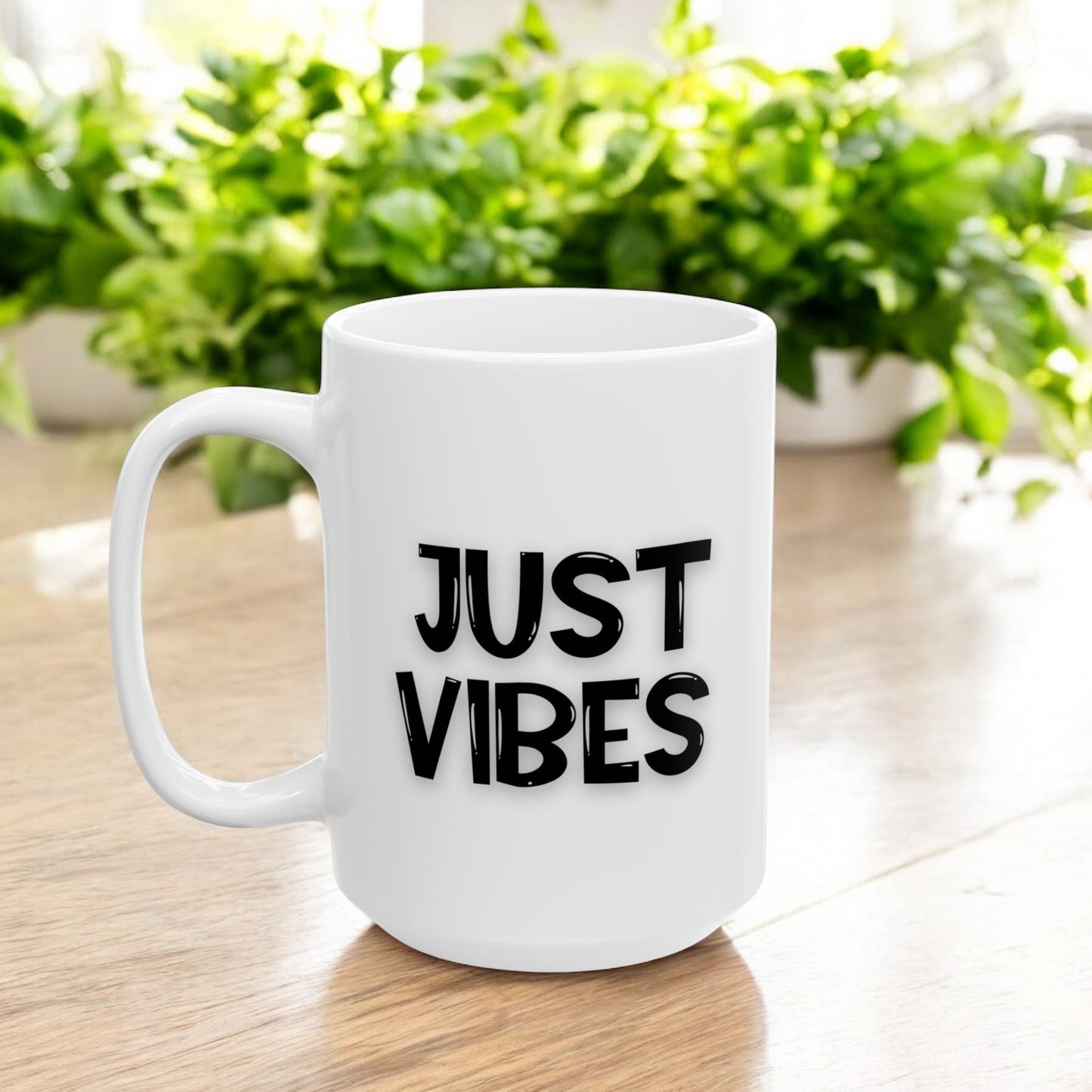 White ceramic 15oz mug with 'Just Vibes' text in bold black font, placed on a wooden table with green plants in the background.