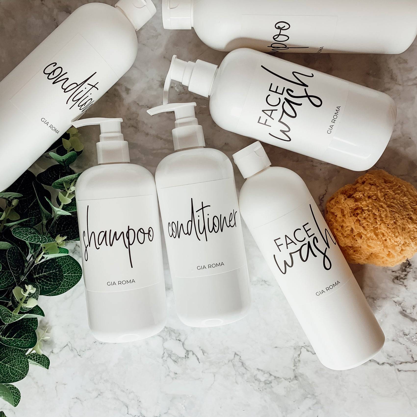 Assorted 16oz white PET pump bottles with custom labels for shampoo, conditioner, and face wash. Reusable dispensers on a marble countertop.