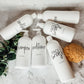 Assorted 16oz white PET pump bottles labeled shampoo, conditioner, and face wash. Reusable dispensers on a marble countertop with greenery.