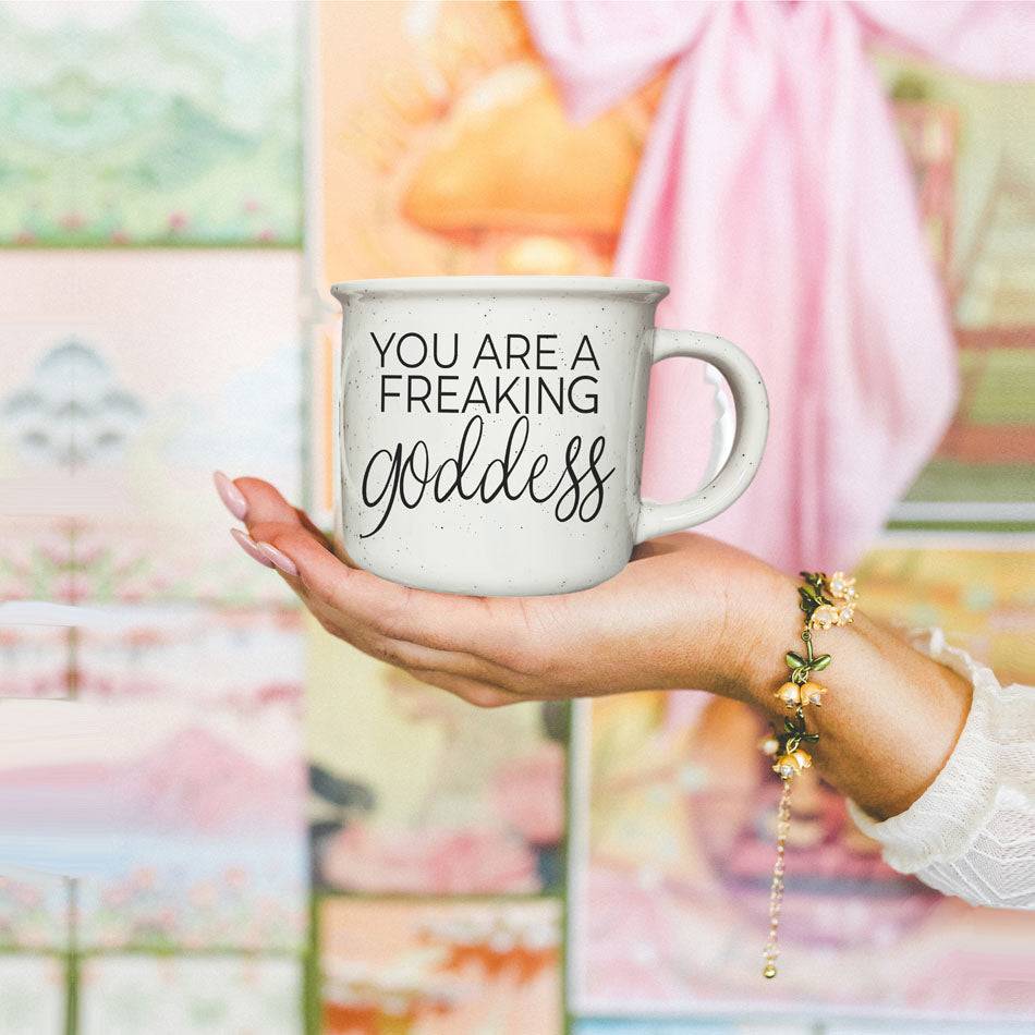 14.5oz ceramic coffee mug with 'You Are a Freaking Goddess' design, held in a hand with a colorful artistic background.